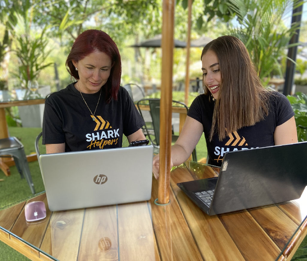Two girls from Shark Helpers talk to each other while checking their laptops, they are experts in hiring Colombian remote experts